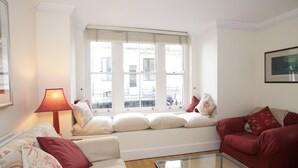 A living room with a sofa, a glass coffee table, a window seat with cushions, and a lamp. - A Place Like Home - Comfortable Apartment in Paddington (London)