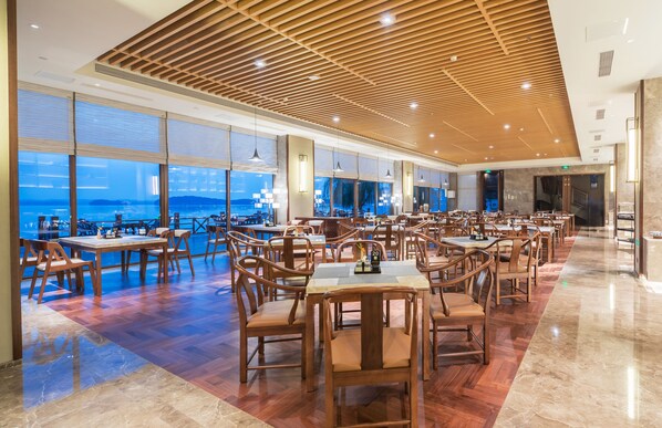 A spacious dining area with wooden tables and chairs, large windows offering a view of the sea, and a wooden ceiling with recessed lighting.