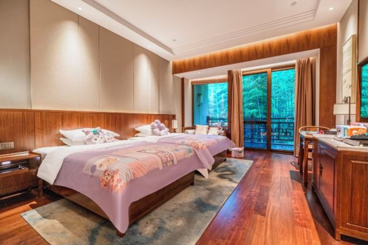 A hotel room with a large bed, wooden paneling, a desk, and a view of trees through the window.