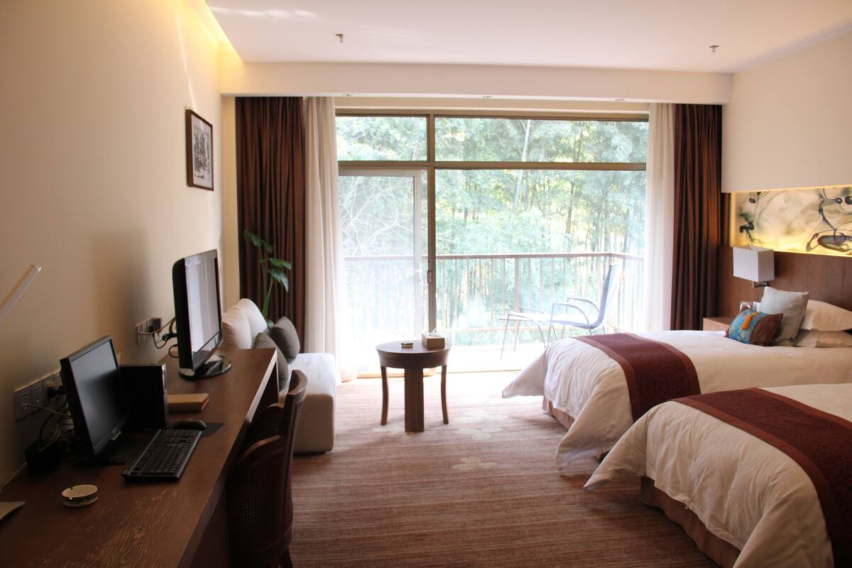 Une chambre d’hôtel équipée d’un bureau, d’un ordinateur, de deux lits, d’une chaise et d’une grande fenêtre donnant sur la forêt.