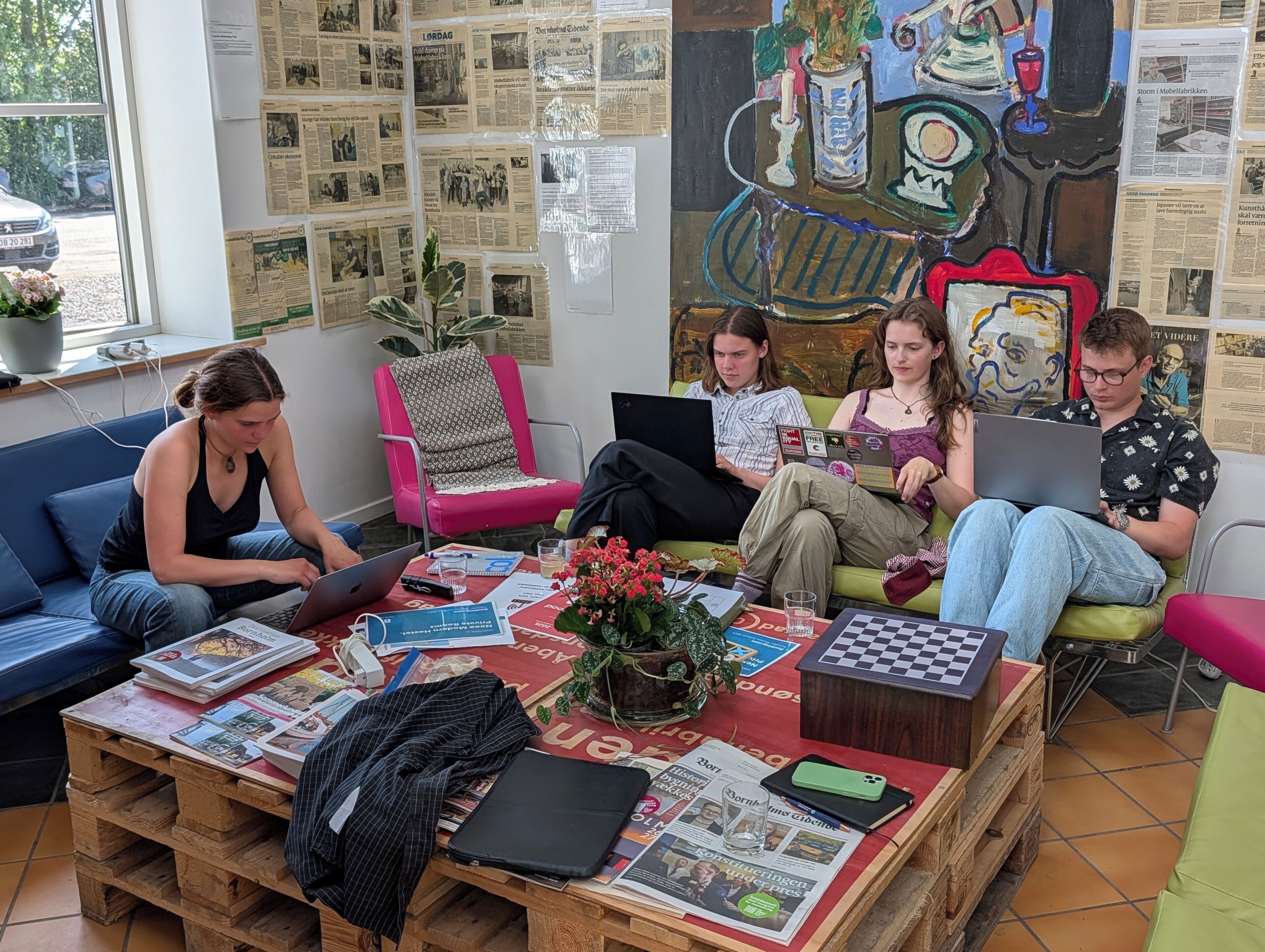 A group of people sitting on a couch and chairs in a room with newspapers on the walls and a table with books and a laptop.