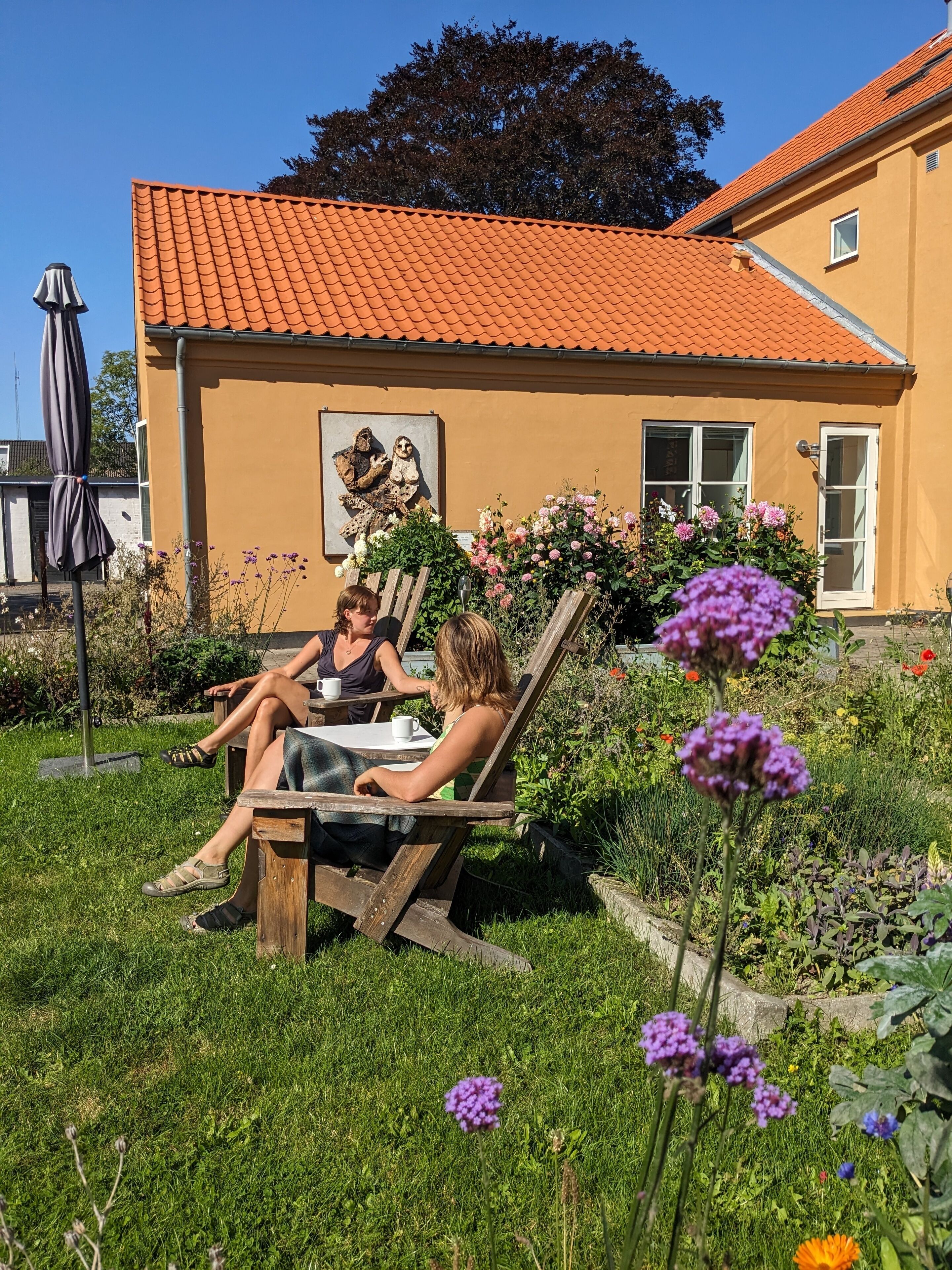 A garden with a wooden bench and a table, surrounded by flowers and a house with a tiled roof.