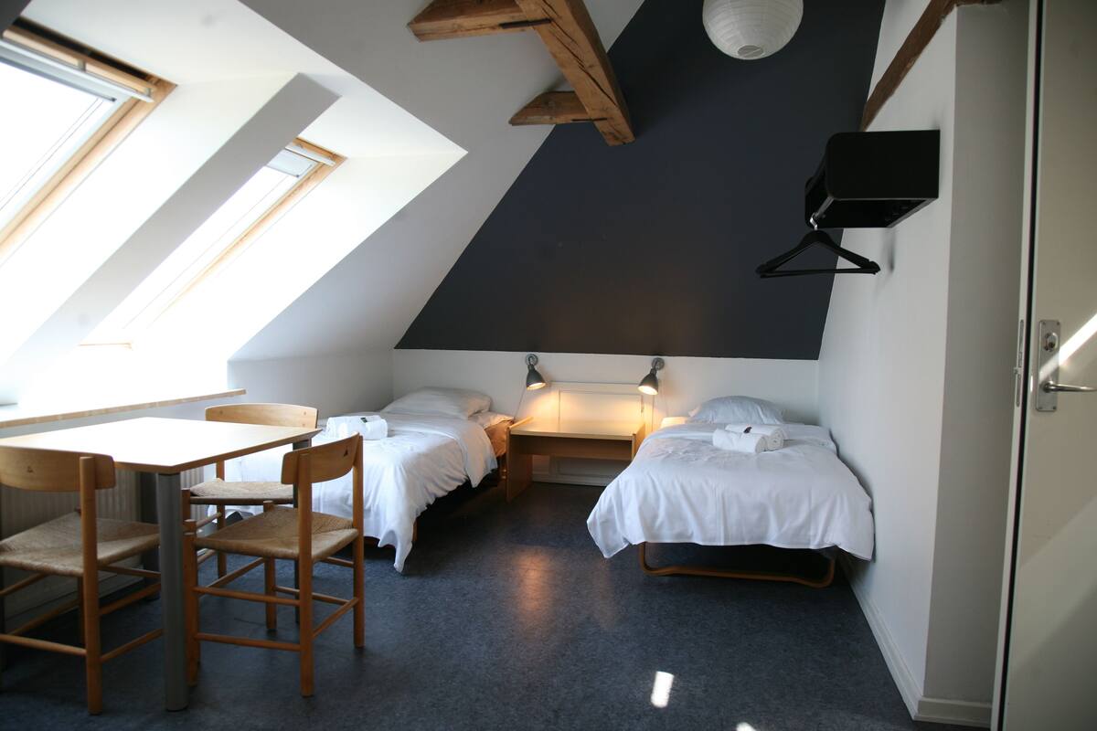 Ein kleiner, gut beleuchteter Raum mit schrägen Wänden, zwei Betten, einem Tisch und Stühlen.