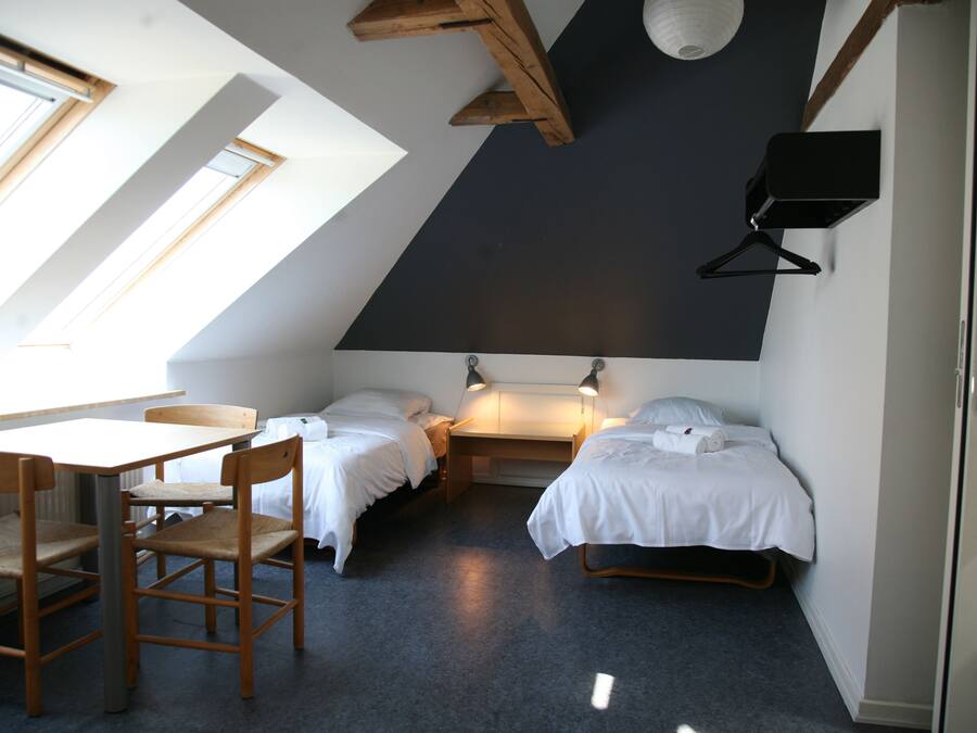 A small, well-lit room with a sloped ceiling, two beds, a table, and chairs.