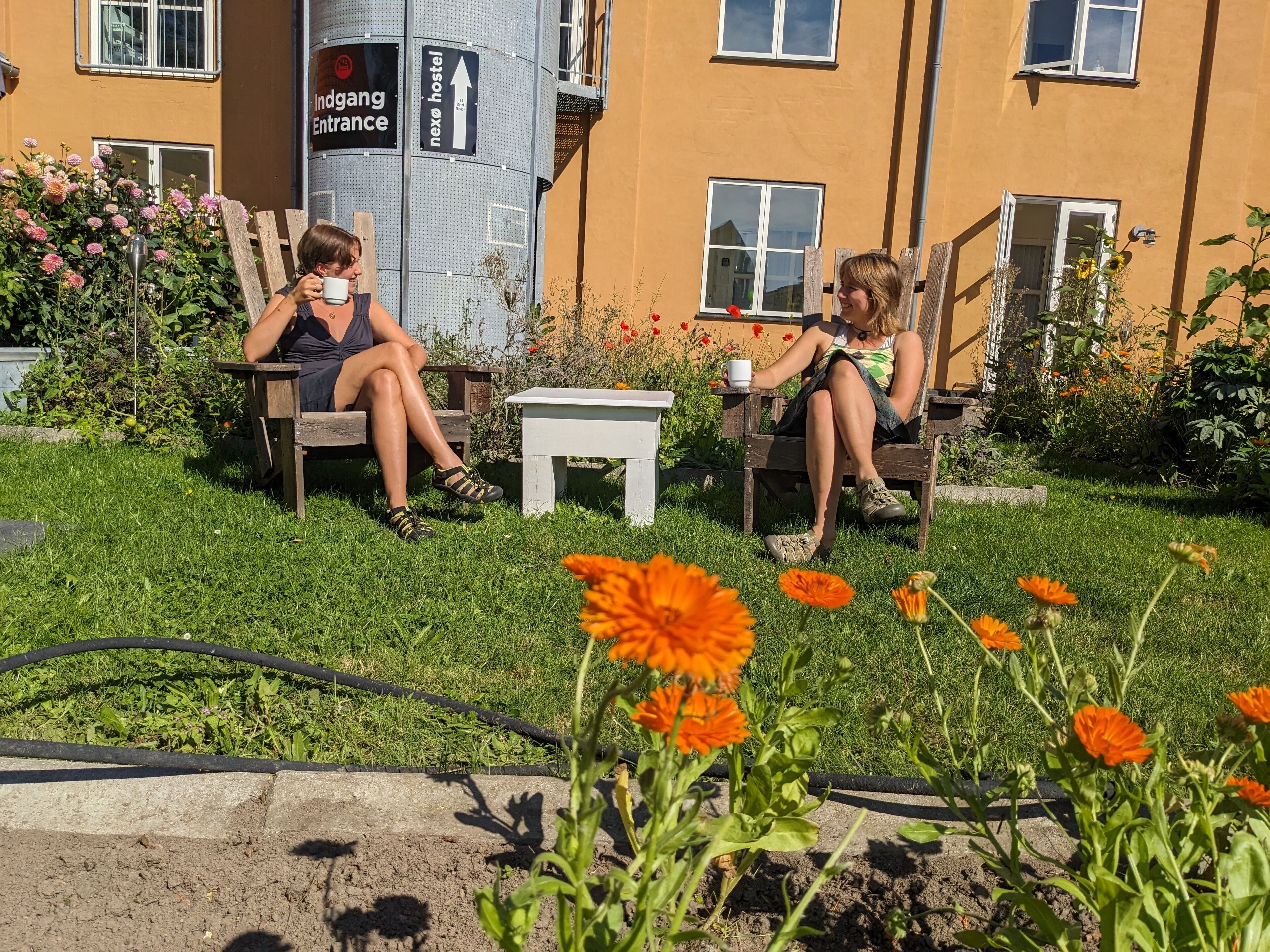 Two people sitting on wooden chairs in a garden area with a building in the background.