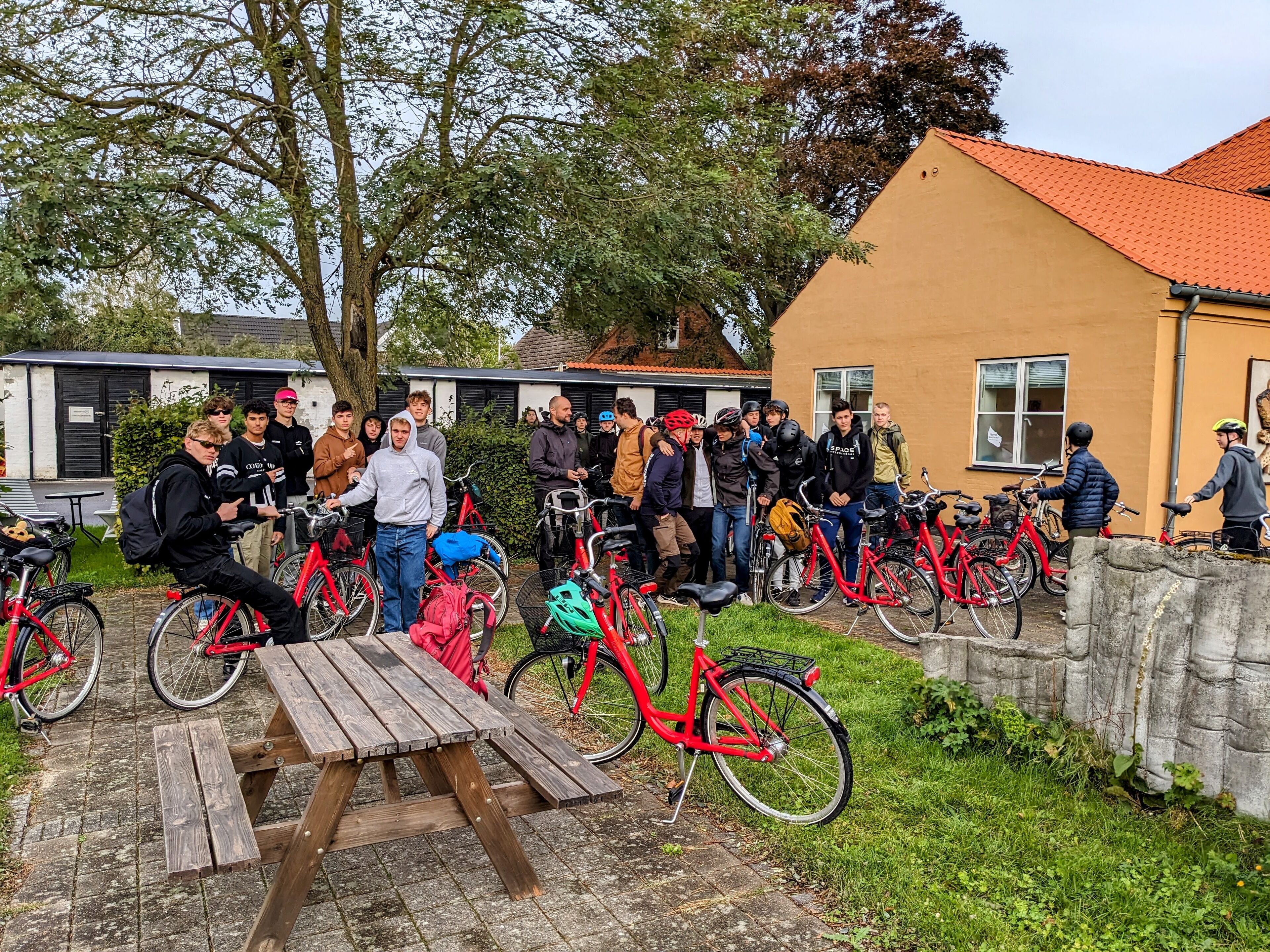 Un gruppo di persone riunite attorno a biciclette e un tavolo da picnic in un ambiente esterno, con un edificio sullo sfondo.