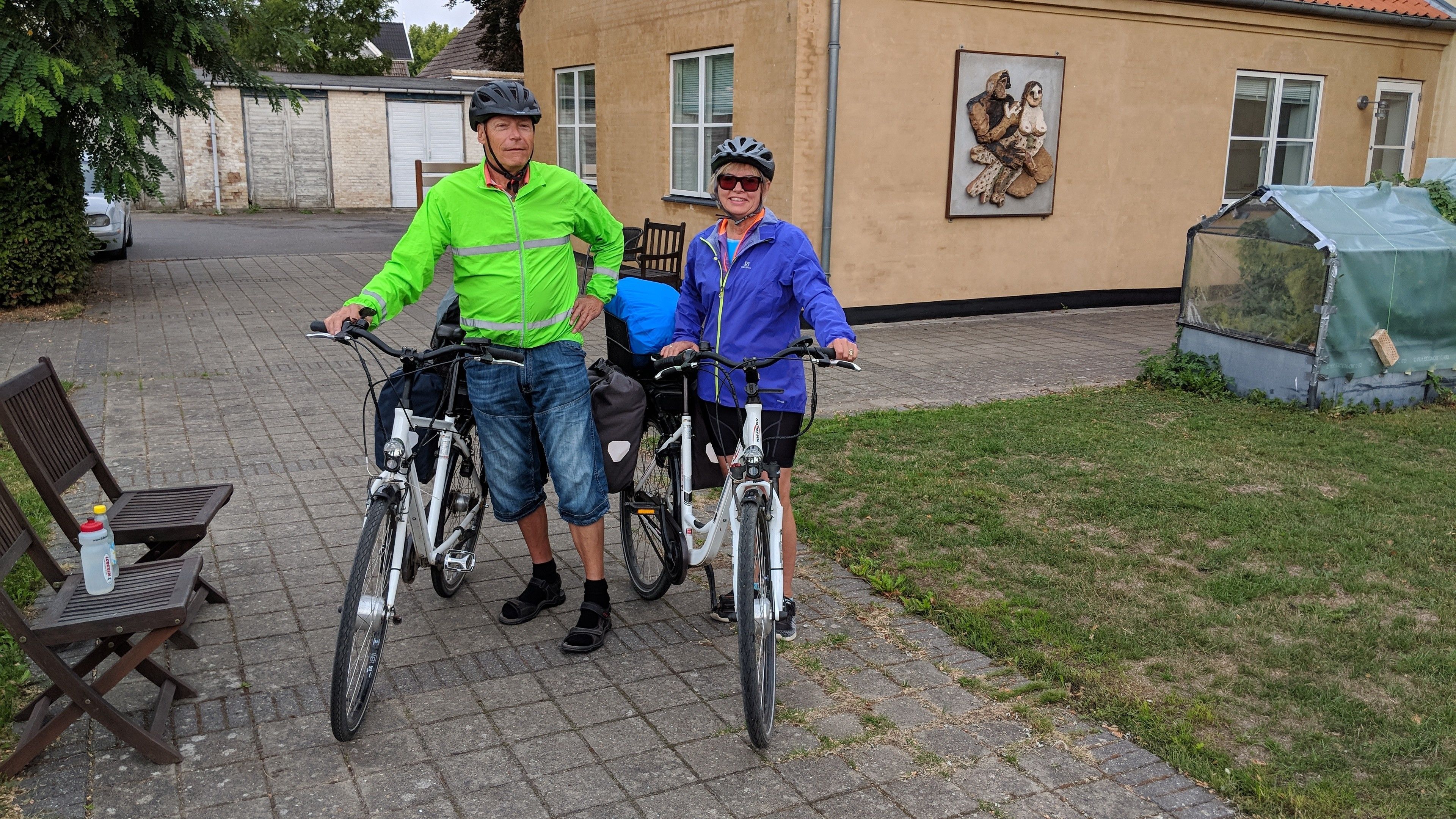 To personer på cykler i en gårdsplads med en bænk og en bygning i baggrunden.
