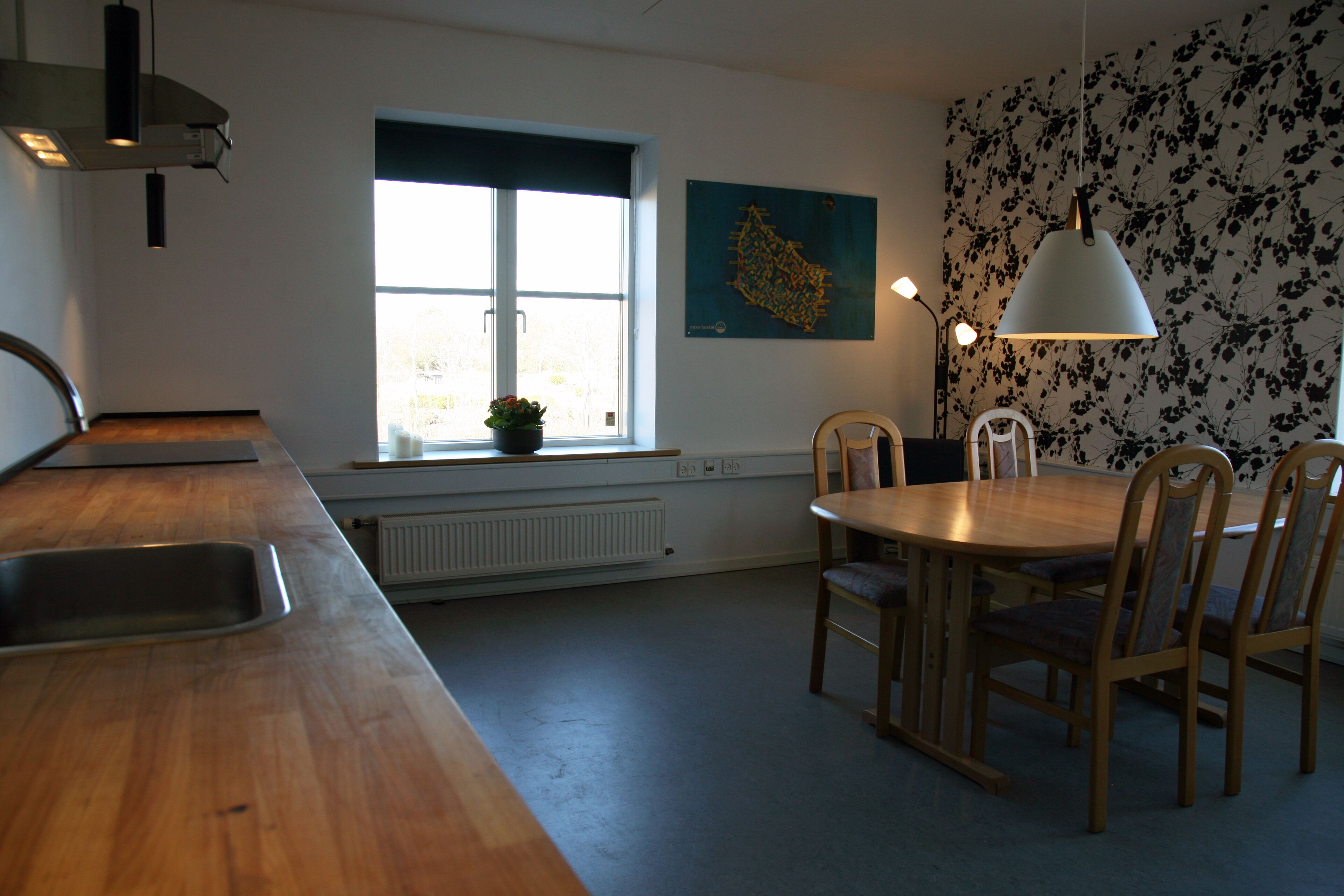 A kitchen with a wooden countertop, a sink, a dining table with chairs, a window with blinds, and a wall with a floral pattern.