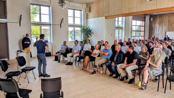 A conference room with a speaker at the front, attendees seated in rows, and a large window providing natural light.