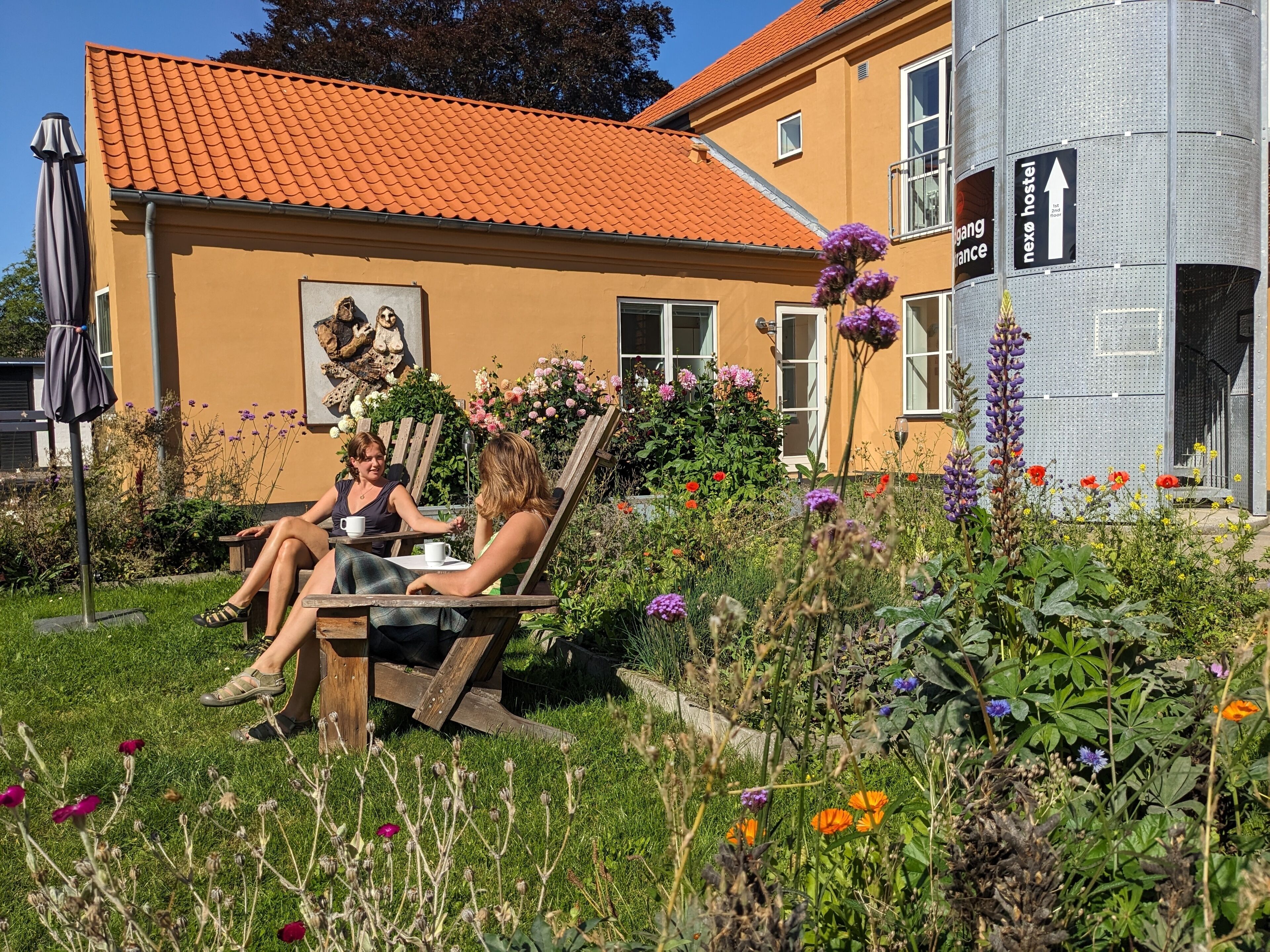 Un jardín con un banco de madera, una mesa y dos personas sentadas. Se observa un edificio con techo rojo y un cartel que dice 'next hostel'.