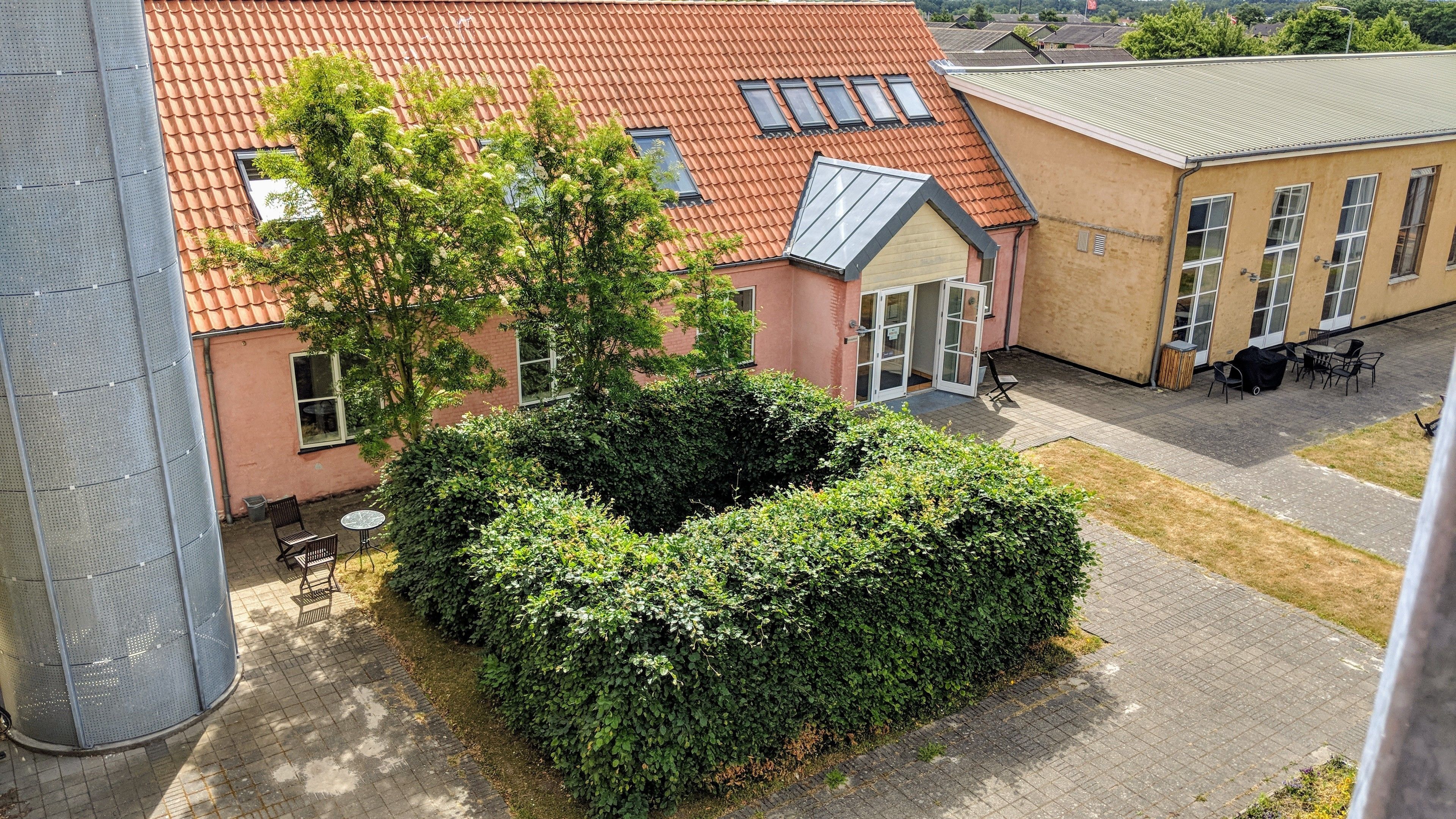 A courtyard with a large hedge, a building with a red-tiled roof, and a seating area with tables and chairs.