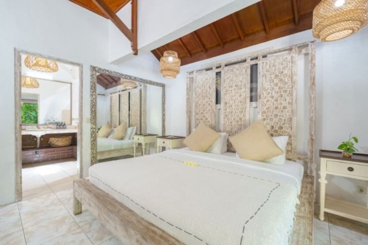 A bedroom with a large bed, a wooden bench, a nightstand, a mirror, and a window with patterned curtains.