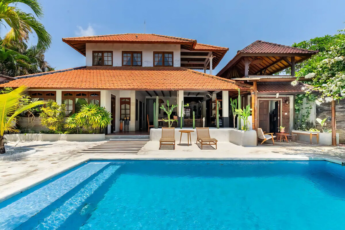 Uma casa de dois andares com piscina, área de estar externa e plantas tropicais.