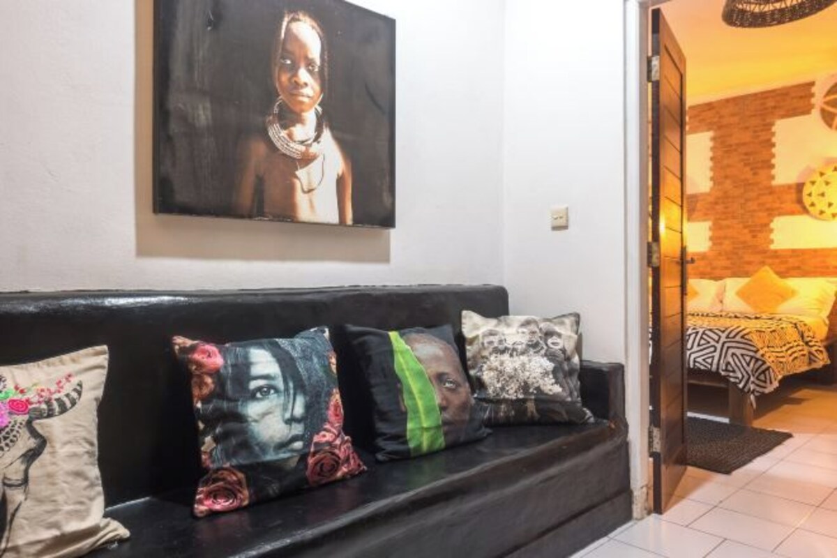 A living room with a black sofa, colorful cushions, a large painting on the wall, and a bed with patterned bedding in the background.