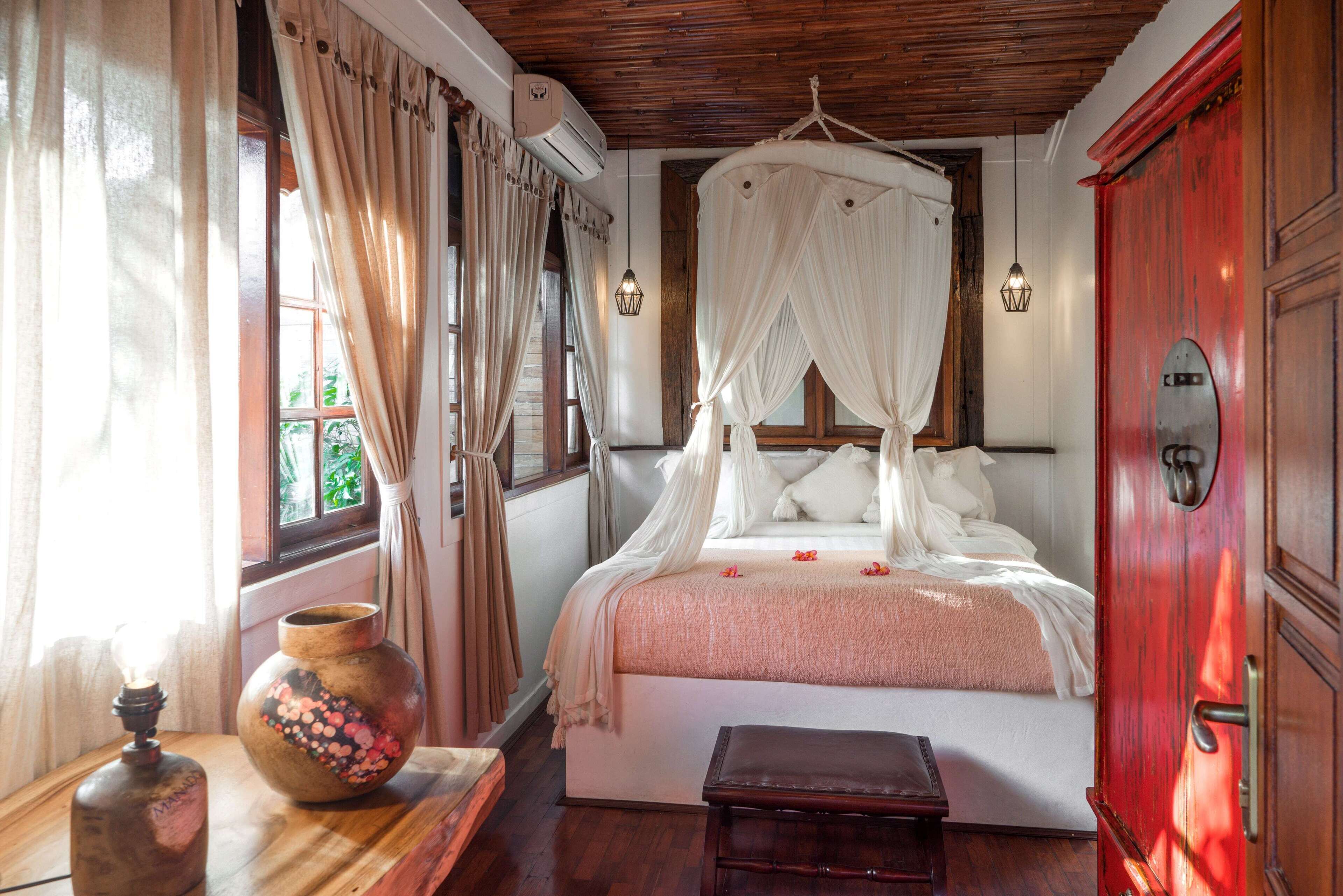 A bedroom with a four-poster bed, a wooden bench, a vase, and a red door.
