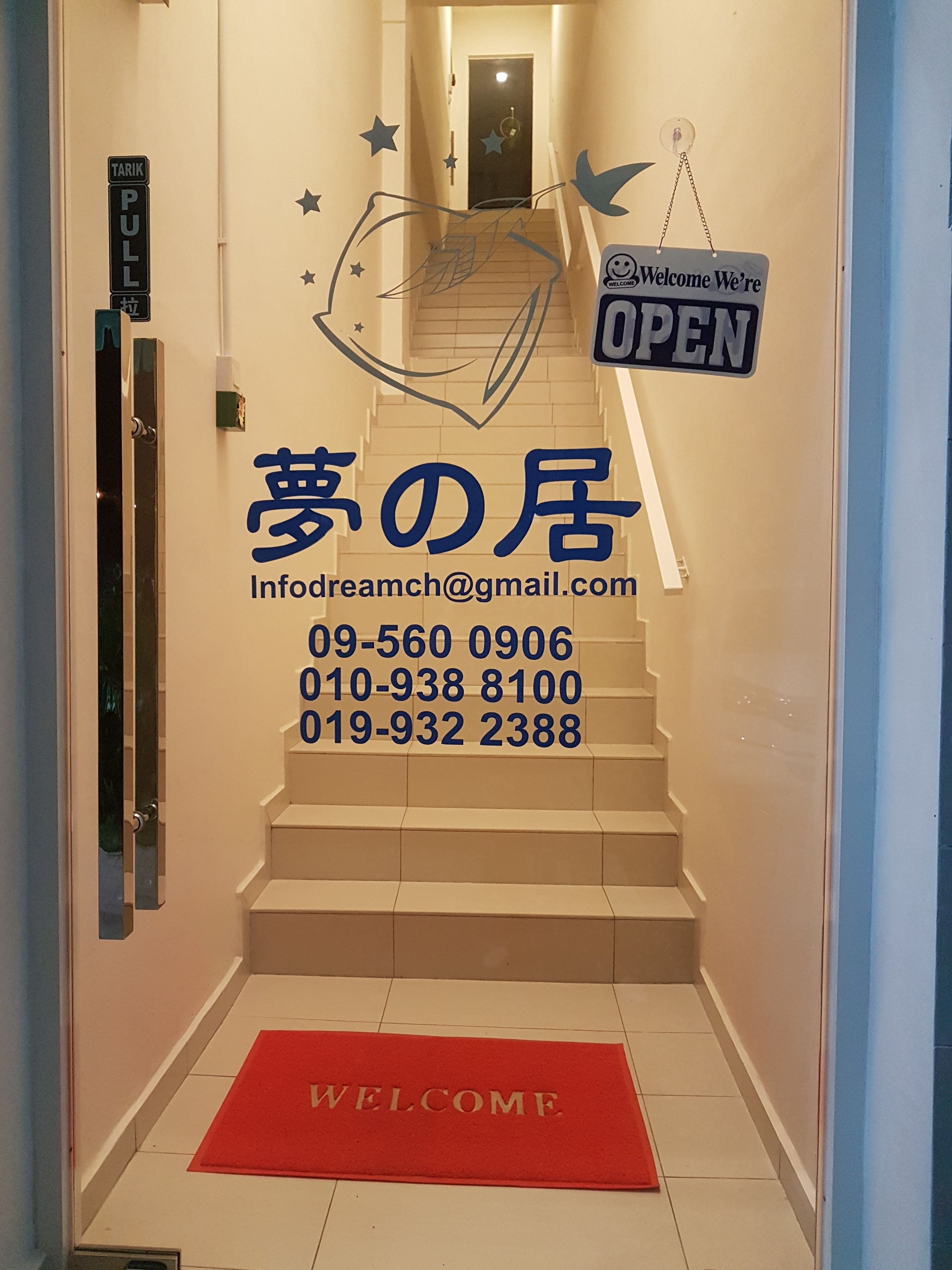 Entrada de un hotel con felpudo rojo, un letrero que dice 'Welcome We're Open' y una escalera que sube al piso superior.