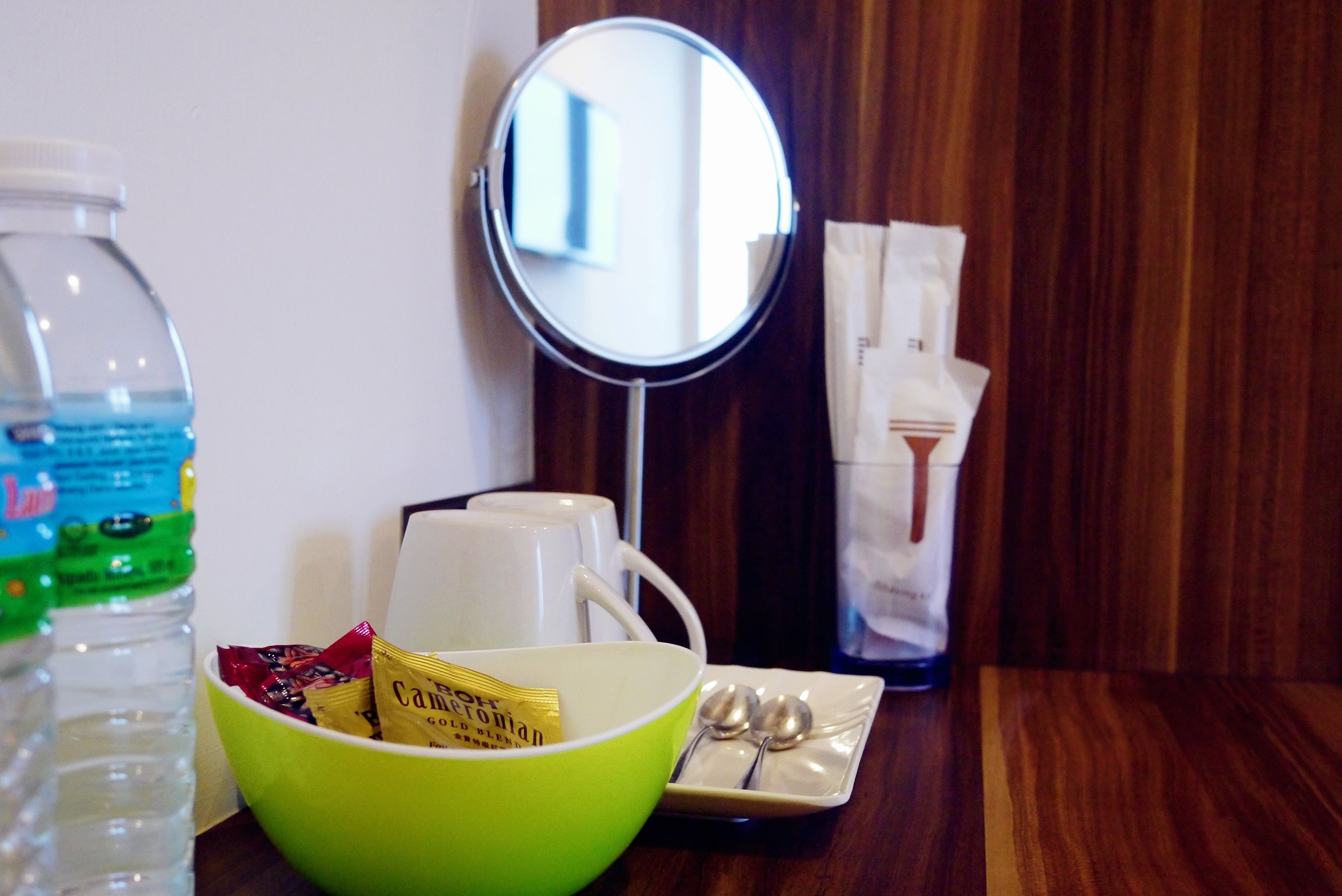 A room with a wooden table, a green bowl with snacks, a white mug, a bottle of water, a mirror, and a tray with two spoons.