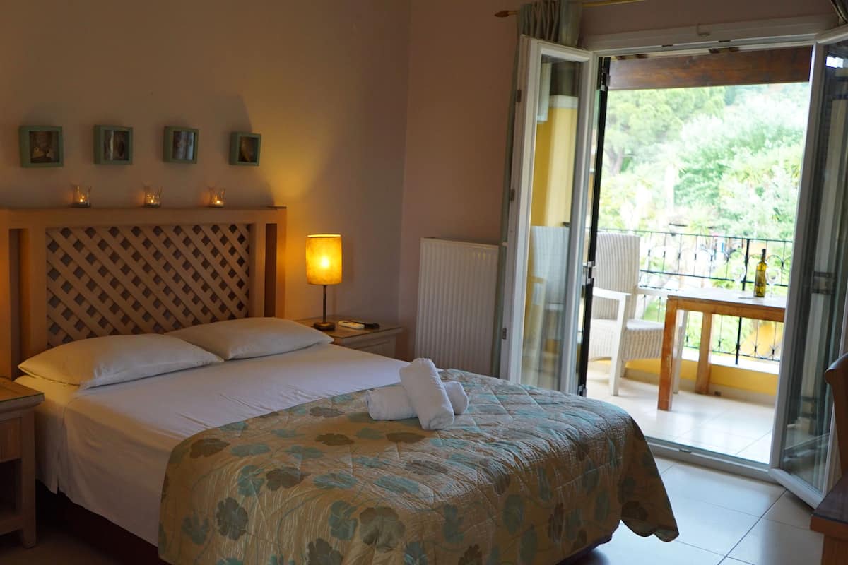 A hotel room with a bed, two bedside lamps, a nightstand, a window with a view of greenery, and a balcony with a table and chairs.