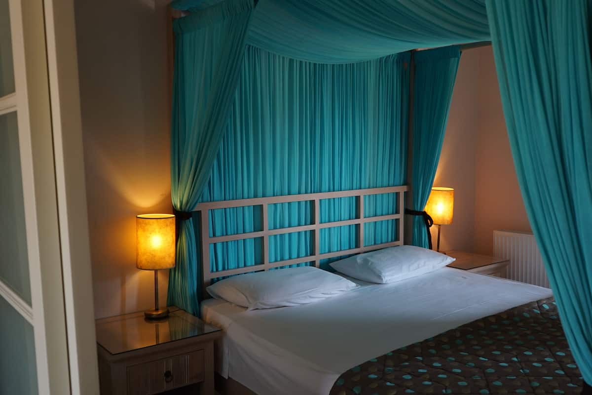 A bed with white bedding, a wooden headboard, and teal curtains. There are two bedside lamps on either side of the bed.