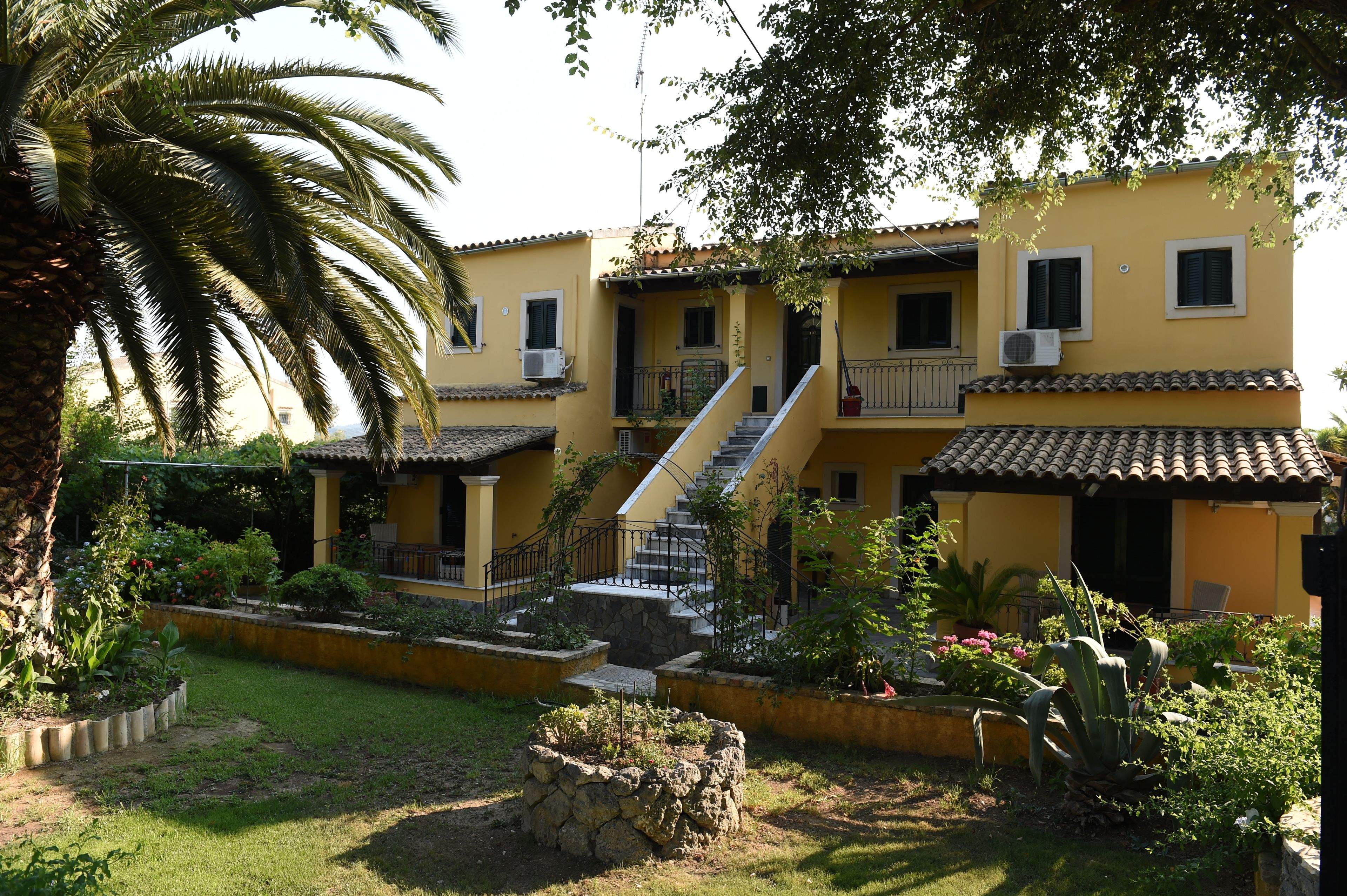 Un edificio amarillo con una escalera que conduce a un balcón, rodeado por un jardín con diversas plantas y una palmera.