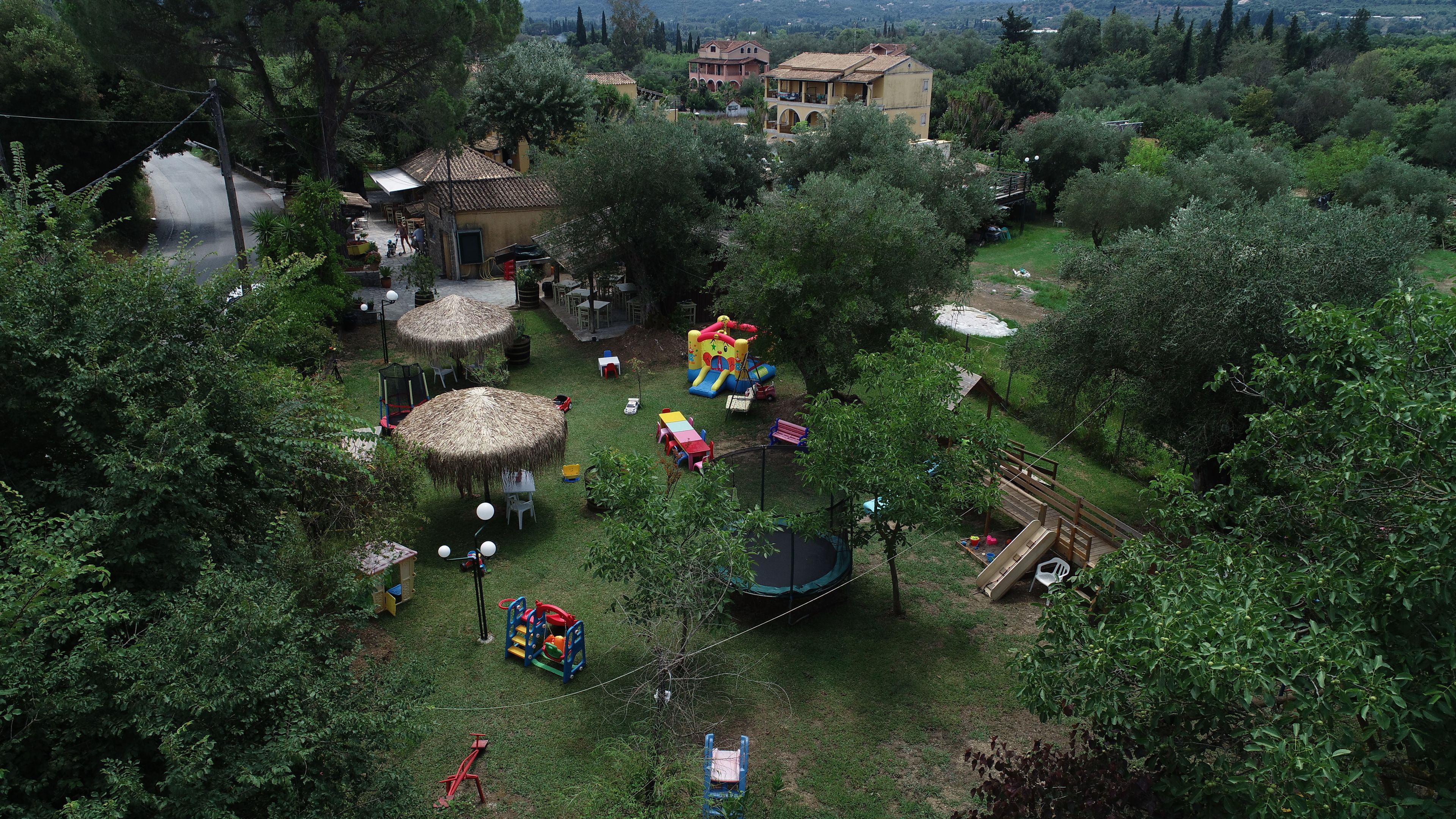 An aerial view of a lush, green park with various play structures, including a slide and a swing set, surrounded by trees and a few buildings in the background.