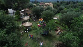 An aerial view of a lush, green park with various play structures, including a slide and a swing set, surrounded by trees and a few buildings in the background.