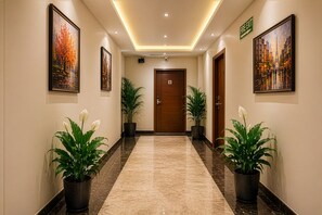 A hallway with a wooden door, potted plants, and framed artwork on the walls. - Hotel Kian by JMD Group (New Delhi)