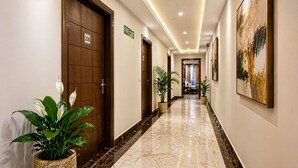 Un pasillo de hotel con piso de mármol, puertas de madera, plantas en macetas y un cuadro en la pared.
