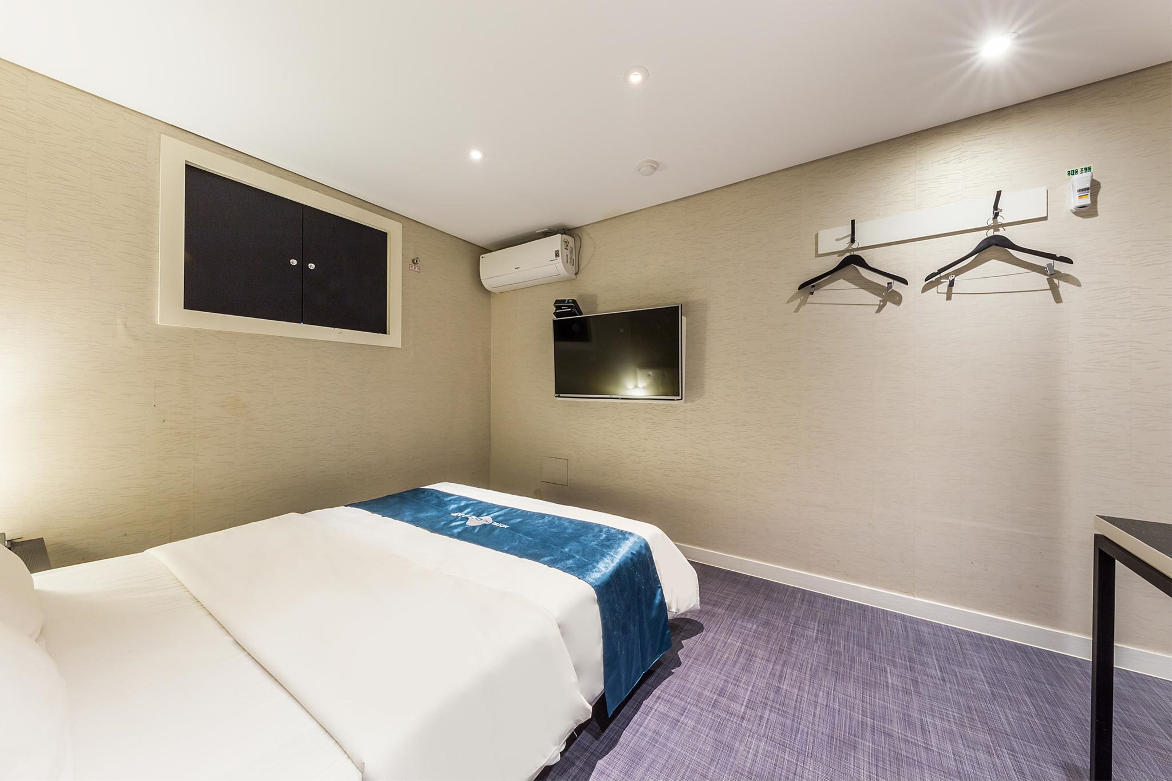 A hotel room with a bed, a flat-screen TV, an air conditioning unit, and a wall-mounted coat rack.