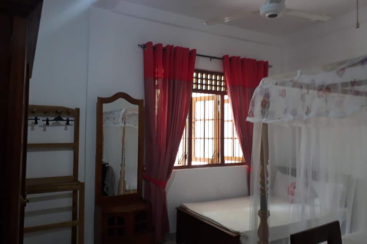 Une chambre à coucher avec un lit en bois, une table de chevet, un miroir et une fenêtre avec des rideaux rouges.