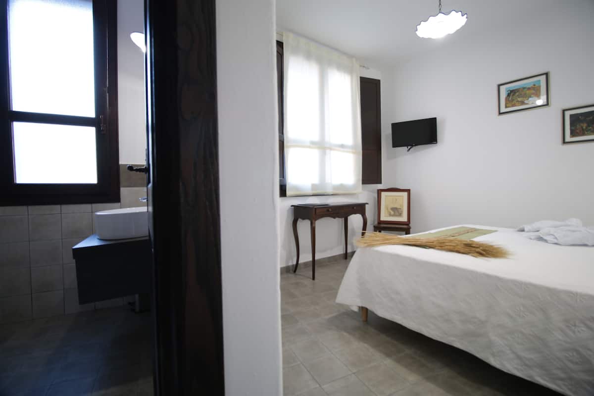Une chambre d’hôtel comprenant un lit, une table de chevet, un miroir, une fenêtre avec des rideaux, un téléviseur fixé au mur et une salle de bain avec un lavabo et des toilettes.