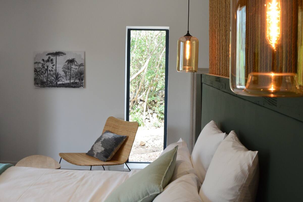 A bedroom with a bed, pillows, a hanging light fixture, a window, and a wall-mounted artwork.