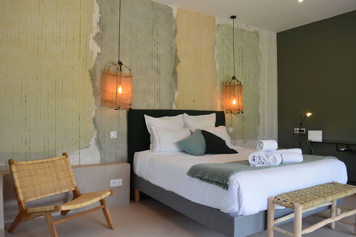 A bedroom with a bed, two wall-mounted lamps, a woven chair, a bench, and a wall with peeling paint.