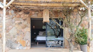 A stone-walled room with a bed, a table, and a hanging light fixture. - Les Petites Maisons (Sotta)