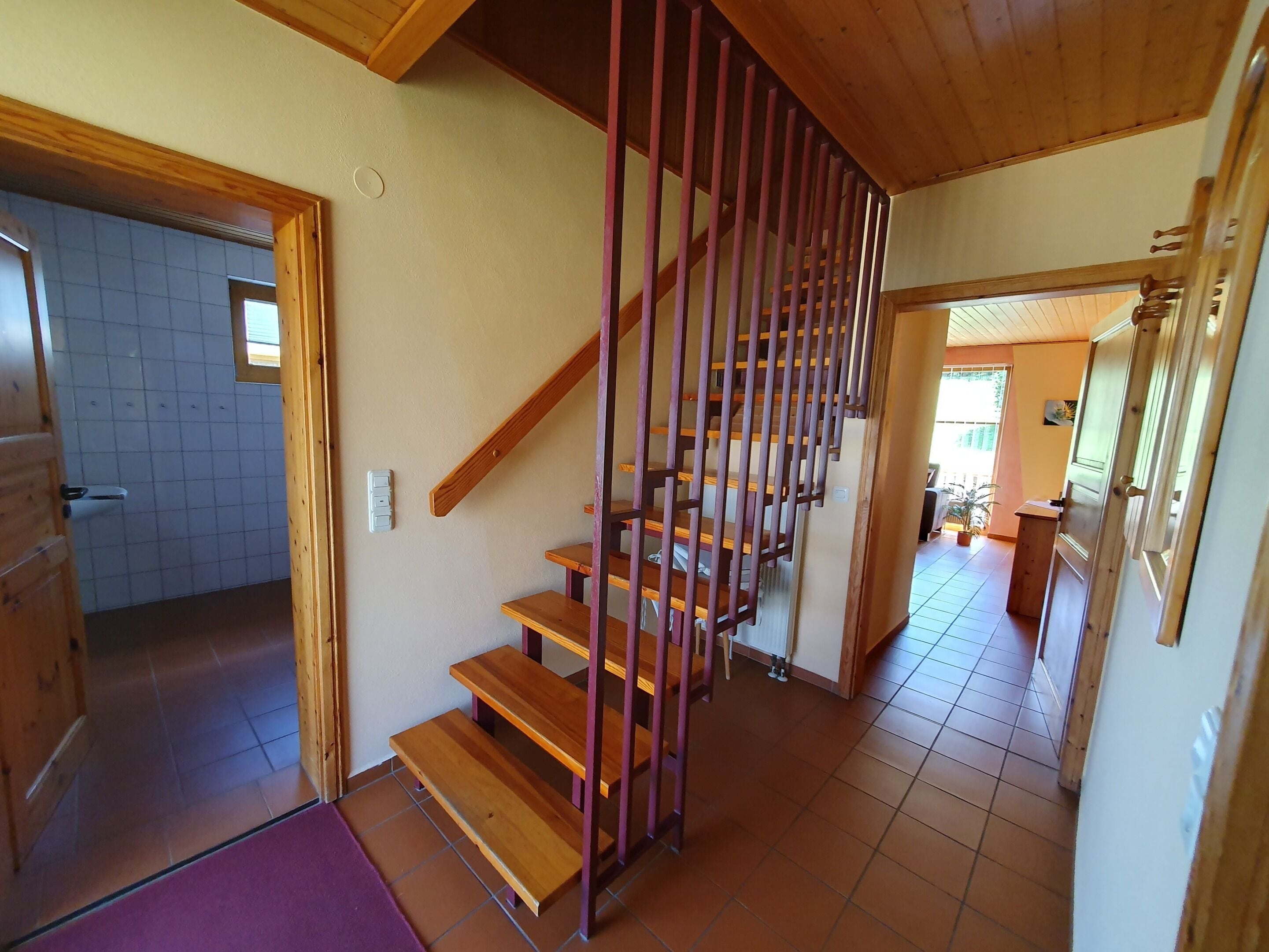 Una escalera de madera con barandilla que conduce a un área del segundo piso con piso de baldosas y una puerta que da a una cocina.
