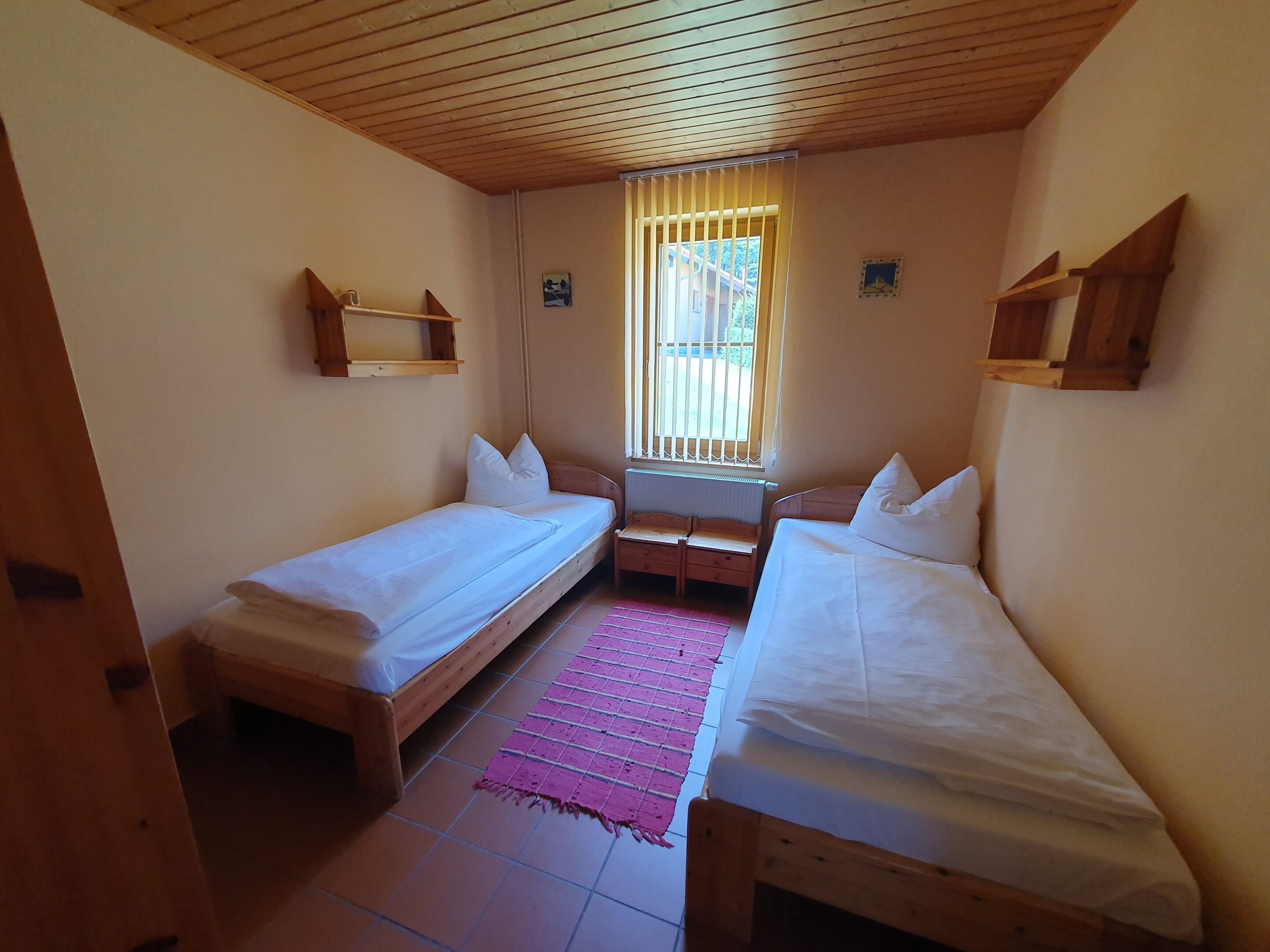 A small, cozy room with two single beds, a window with bars, and a wooden ceiling.