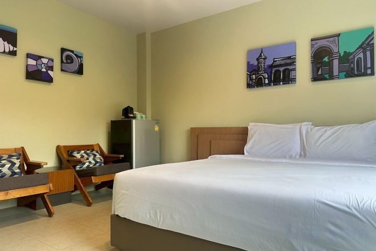 A hotel room with a bed, a desk, and chairs. There are wall art pieces and a refrigerator.