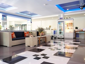 A modern lobby with a checkered floor, a sofa with floral cushions, a coffee table, and a refrigerator stocked with beverages.