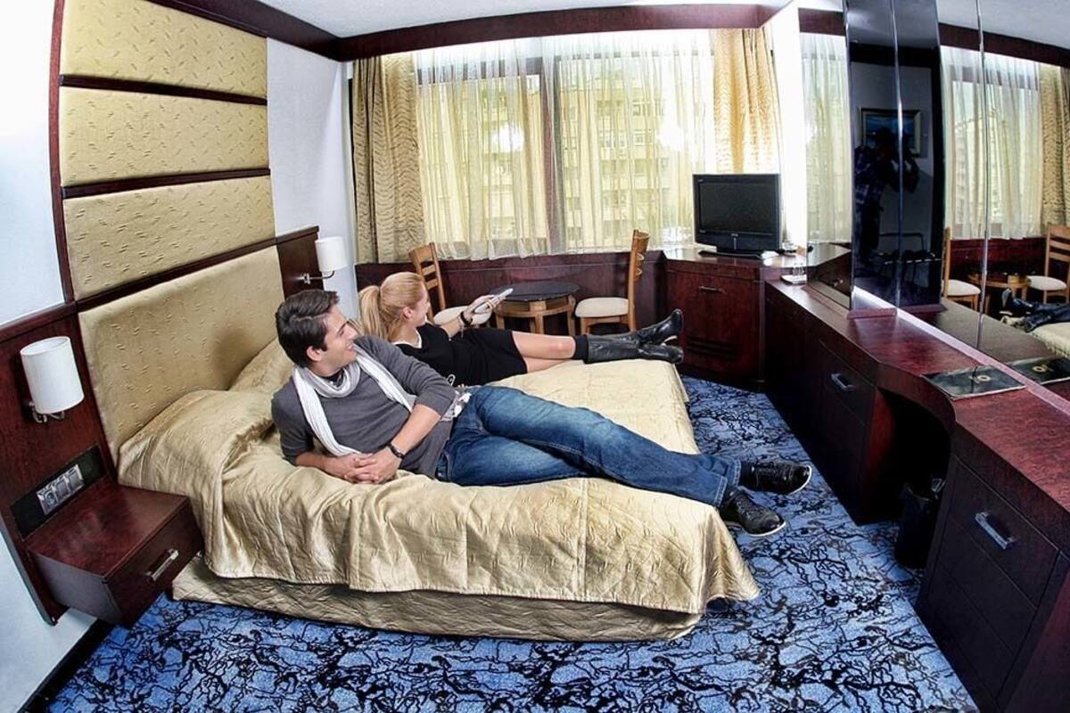 Quarto de hotel com cama, escrivaninha, televisão e espelho.