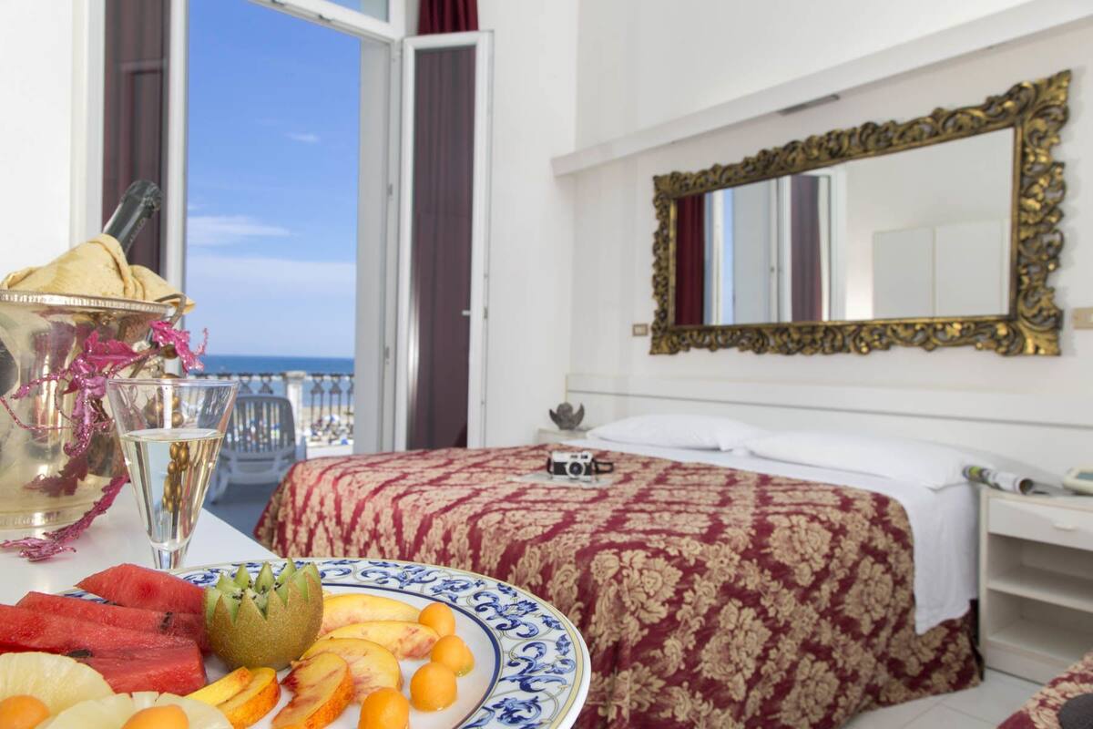 Ein Hotelzimmer mit Bett, einem Nachttisch mit Kamera, einer dekorativen Platte mit Früchten und Meerblick durch eine offene Tür.