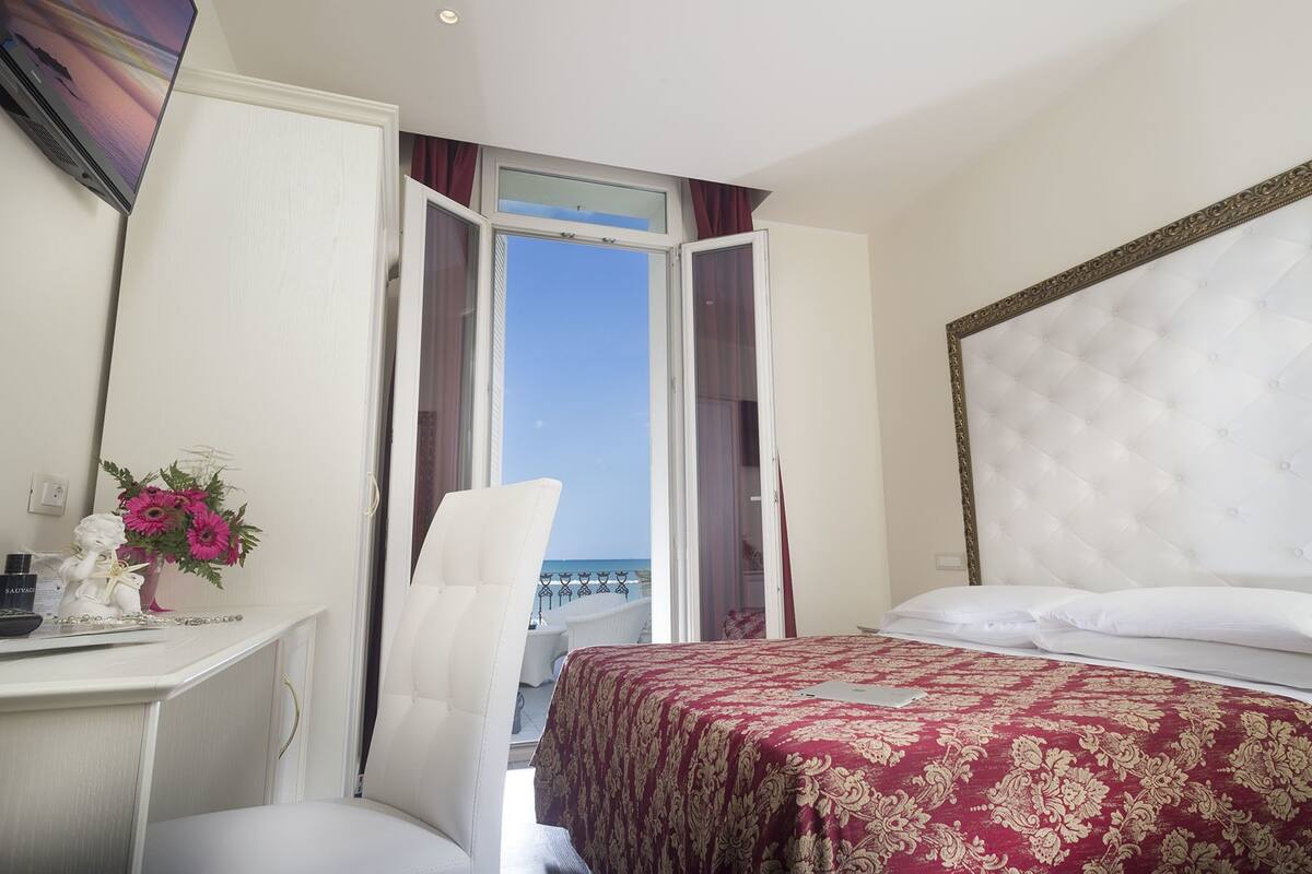 Ein Hotelzimmer mit einem Bett, einem Schreibtisch und einem Stuhl. Ein Fenster bietet Blick auf das Meer.