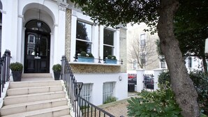 A two-story building with a black front door, white trim, and a small balcony with potted plants. - A Place Like Home - Elegant flat in South Kensington (London)