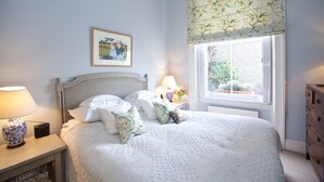 A bedroom with a bed, a nightstand, a lamp, a dresser, a window with floral curtains, and a framed picture on the wall. - A Place Like Home - Elegant flat in South Kensington (London)