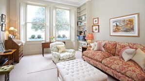 A living room with a green armchair, a patterned sofa, a white ottoman, a wooden desk, and a bookshelf. - A Place Like Home - Elegant flat in South Kensington (London)