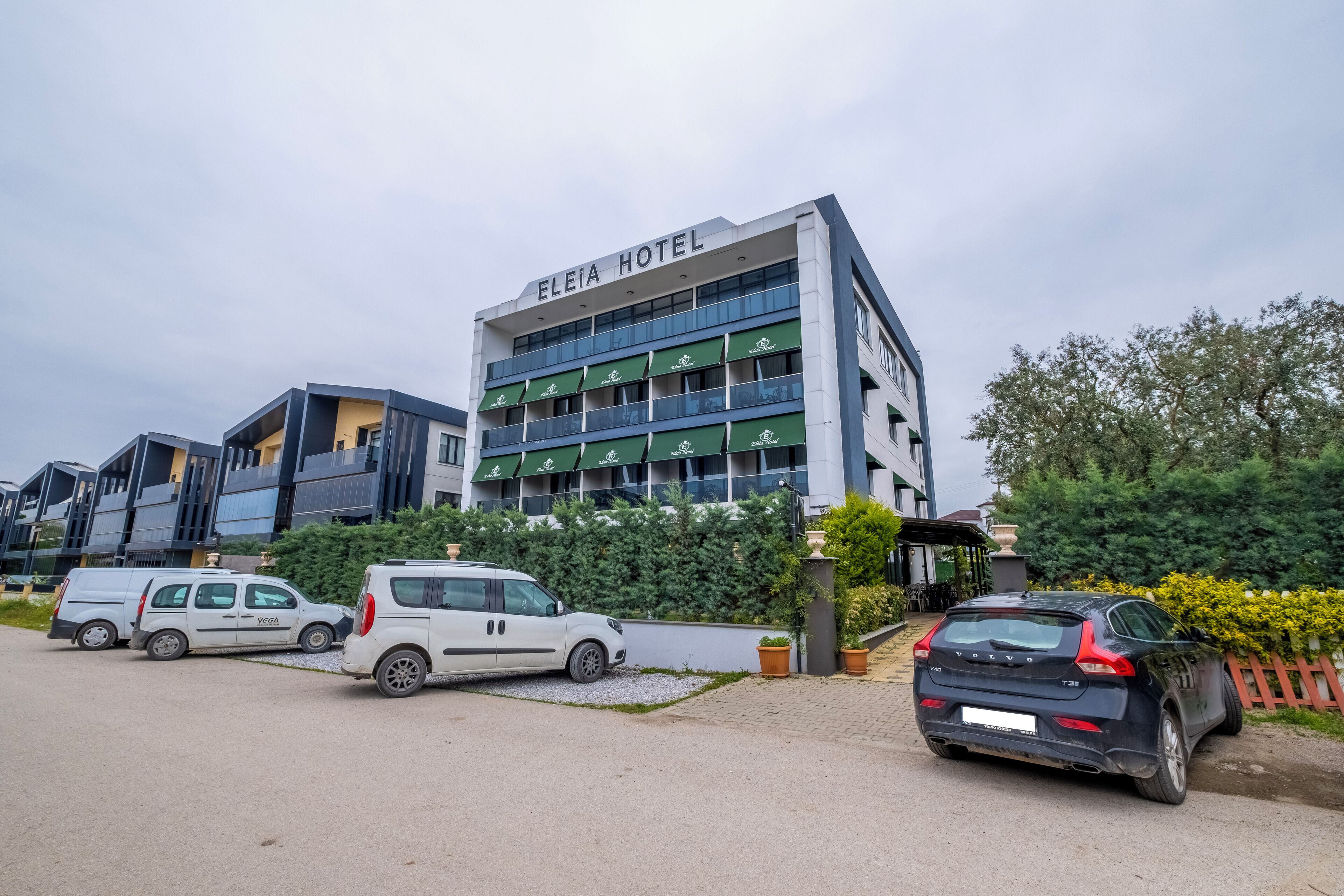 A hotel with a modern design, featuring a prominent sign "ELEIA HOTEL" and multiple balconies. There are several parked vehicles, including a white van and a black car.