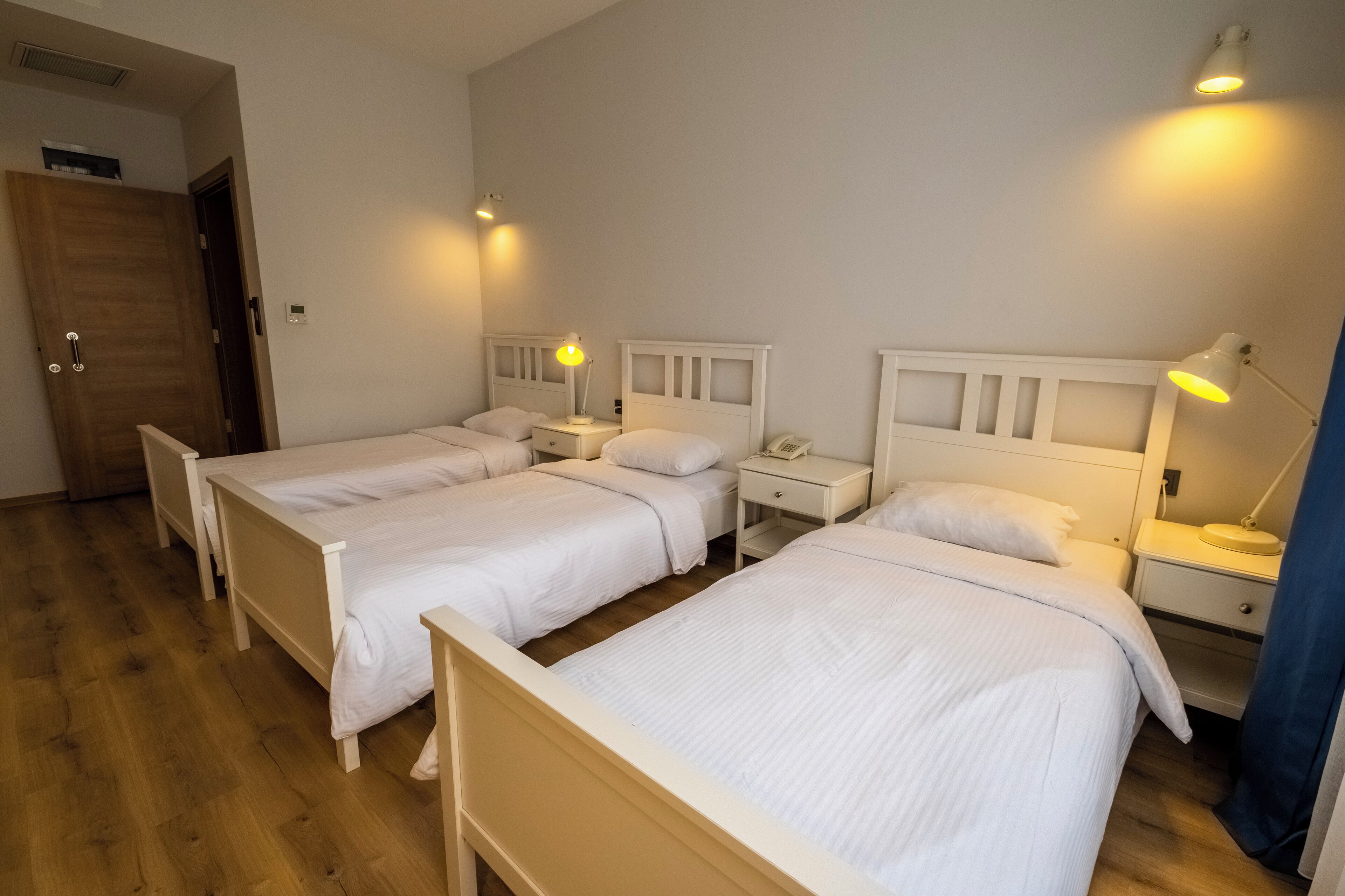 Ein Hotelzimmer mit drei Einzelbetten, Holz-Kopfteilen, Nachttischen mit Lampen und Holzboden.