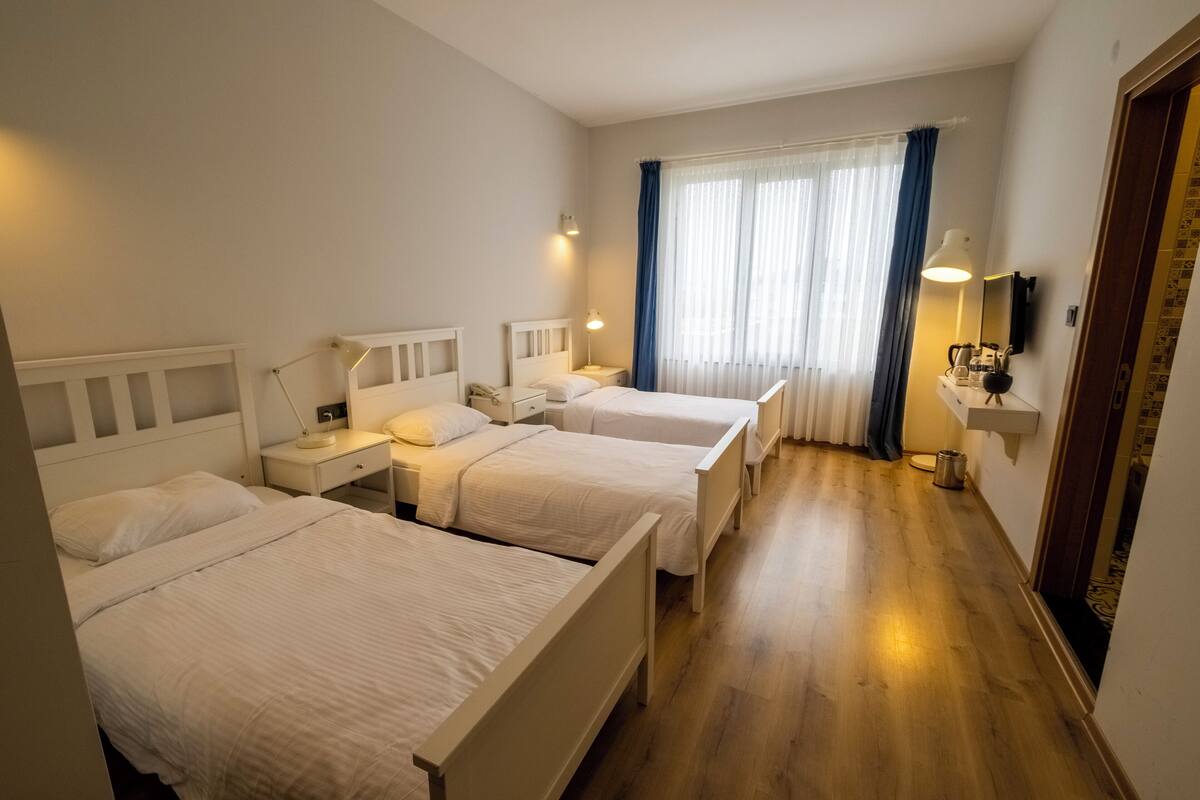 A hotel room with three single beds, a bedside table with a lamp, a small desk with a chair, a window with curtains, and a wall-mounted TV.