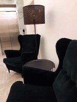Uma área de estar com dois poltronas de cor escura e uma mesa redonda pequena, um abajur de chão alto com cúpula perfurada, e um elevador visível ao fundo.