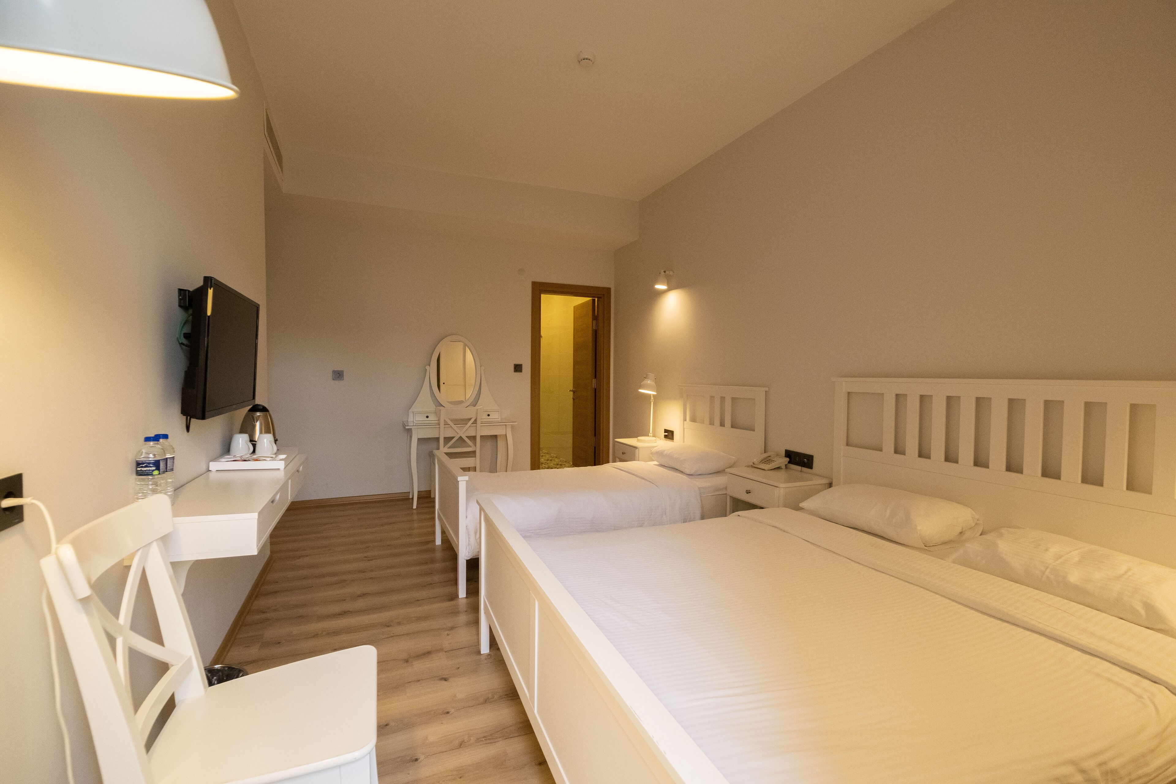 Habitación de hotel con dos camas, un escritorio con silla, un espejo, un televisor y una mesita de noche con lámpara.