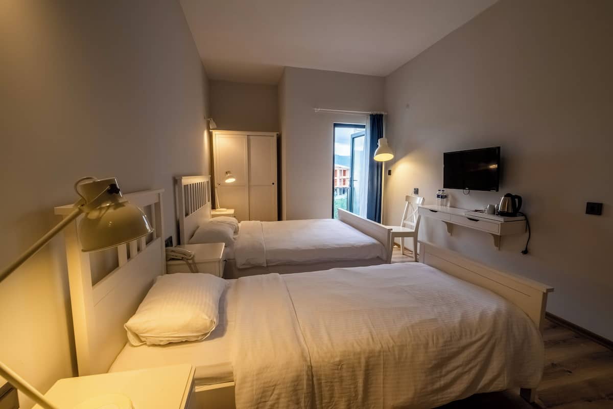 A hotel room with two beds, a desk, a chair, a TV, and a window with a view of buildings.